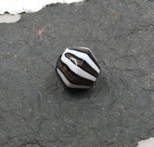 A white, black and metallic striped large bicone glass bead sitting on dark grey handmade paper.