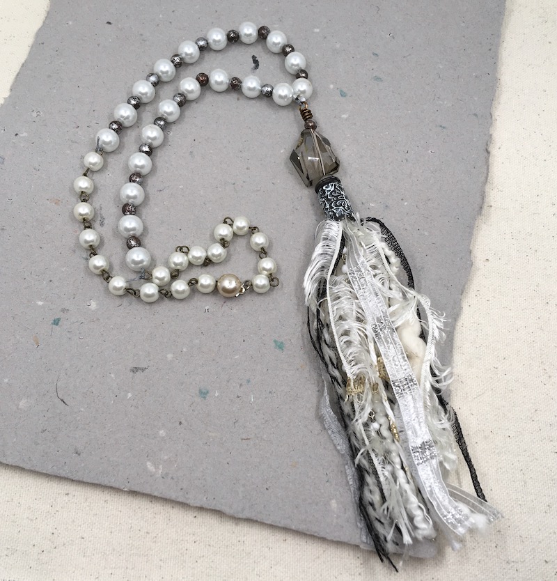 A necklace with white pearls, rustic metal spacers, a large grey crystal, a back and white tassel cap over white and black fibers forming a tassel.