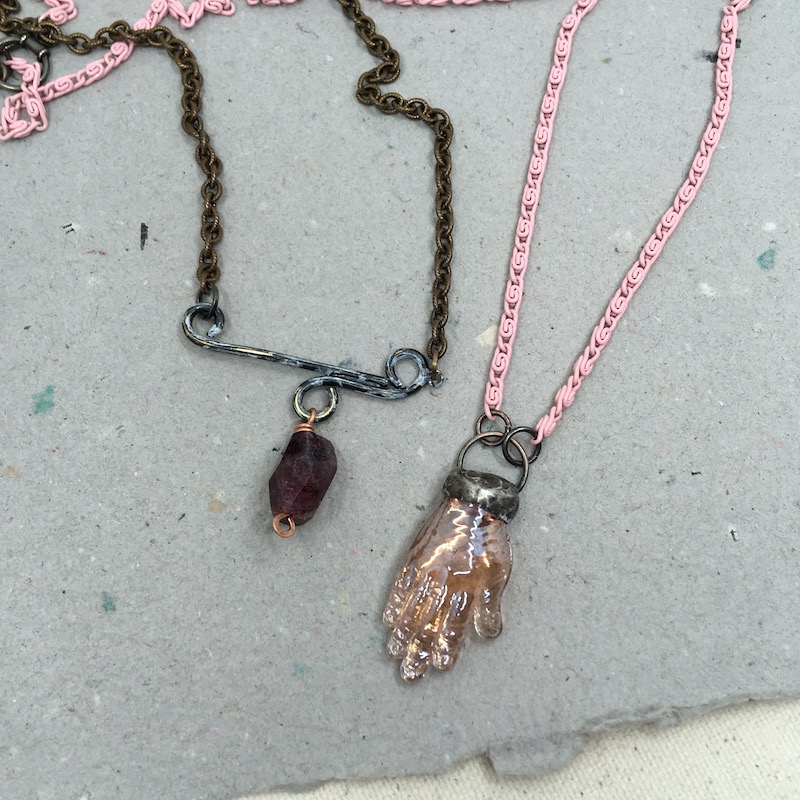 Two necklaces. One with pink glass hand on pink chain. The other a dark rose bead on a metal bar with brass chain.