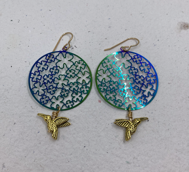 Earrings with round blue and green metal charms with cutout four leaf clover design and brass hummingbird charms. 