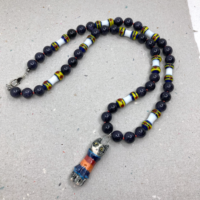 Necklace with cat wearing a sweater pendant, blue goldstone round beads, and manik manik barbells in a white, blue, yellow and red design. 